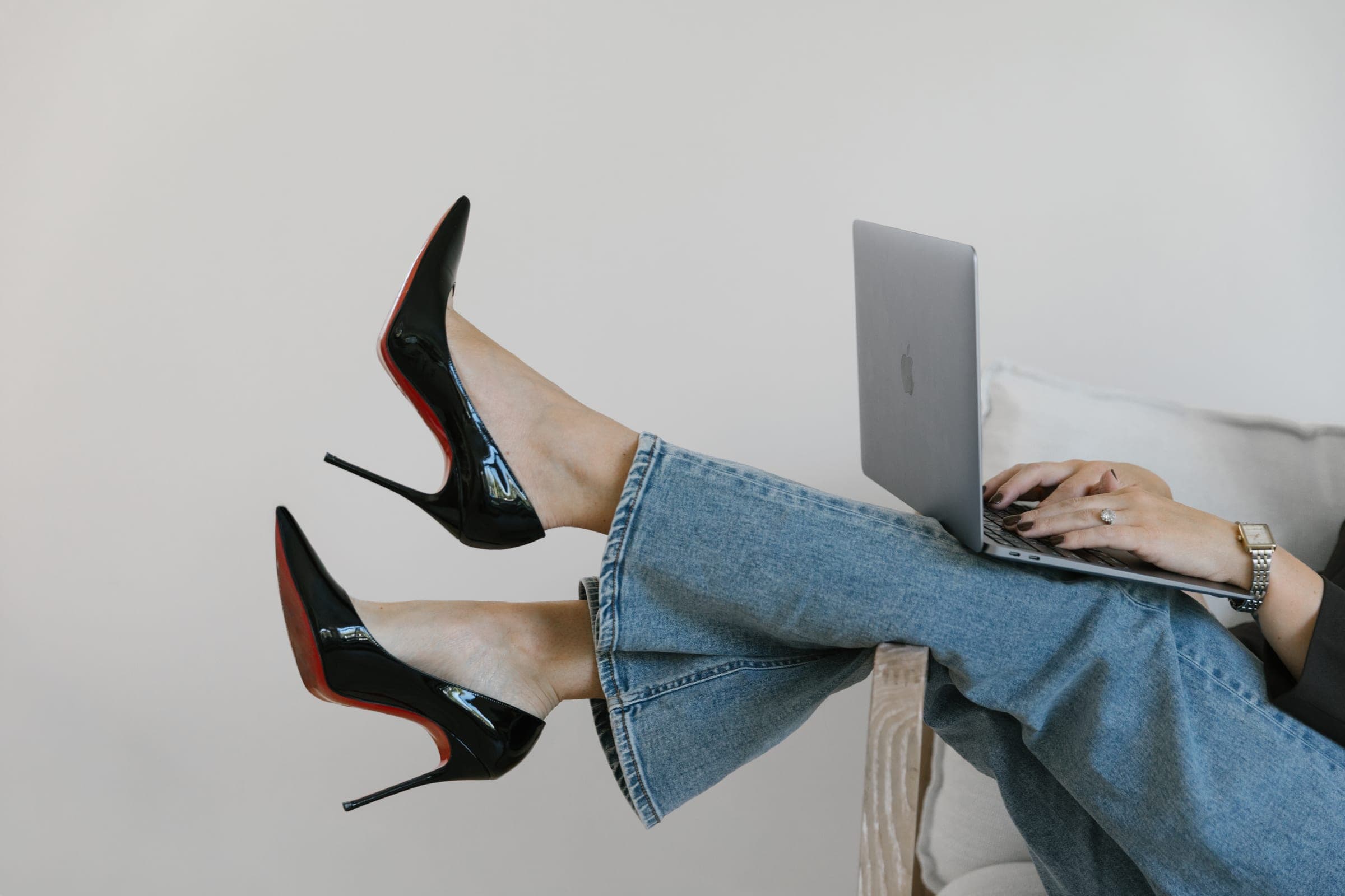 Brittany Saari in jeans and black patent heels, feet up on a chair arm, working on a laptop