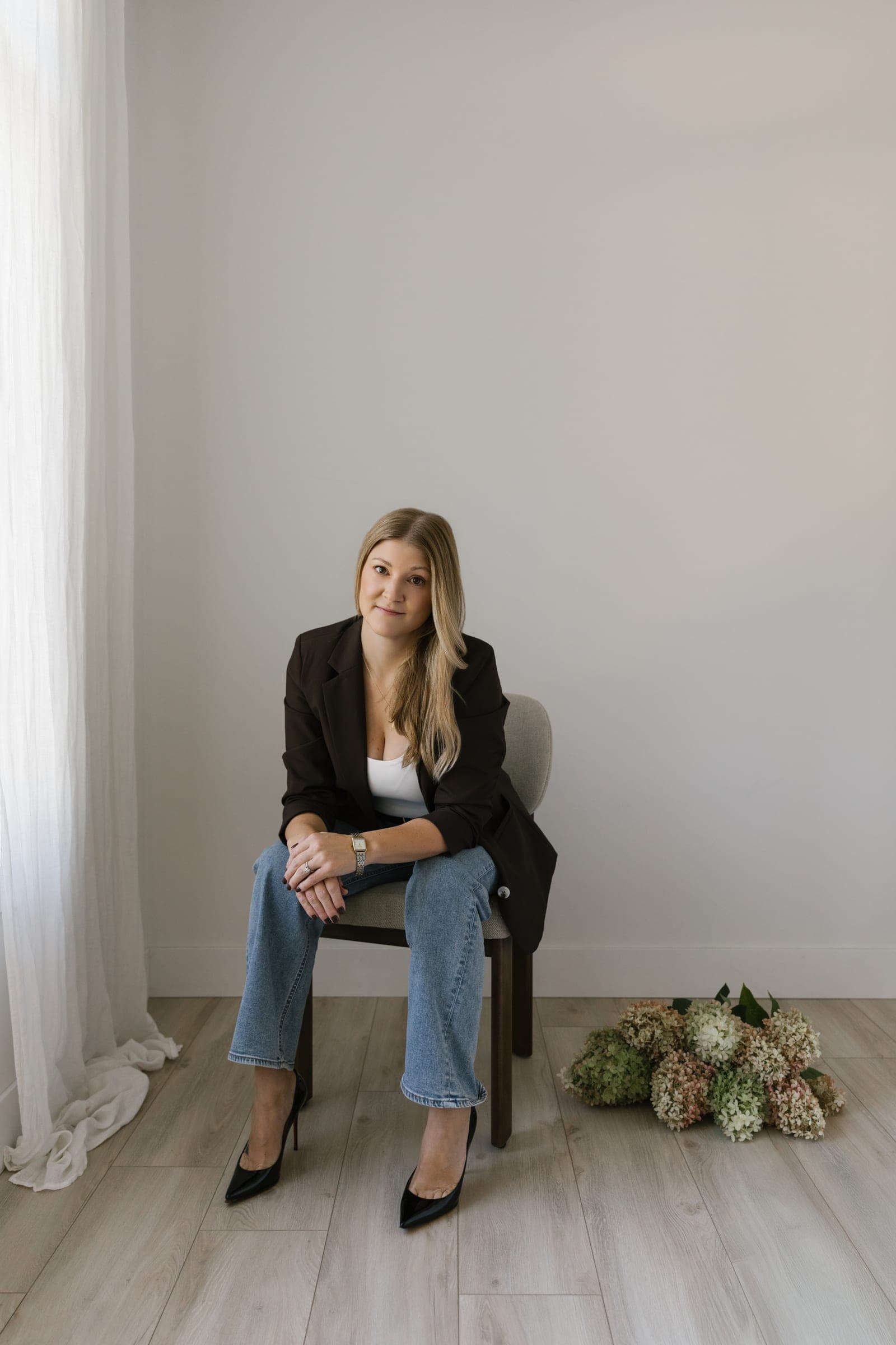 Brittany Saari, owner and principal planner of Walker & West, seated on a chair in a dark blazer and jeans beside a bundle of hydrangeas