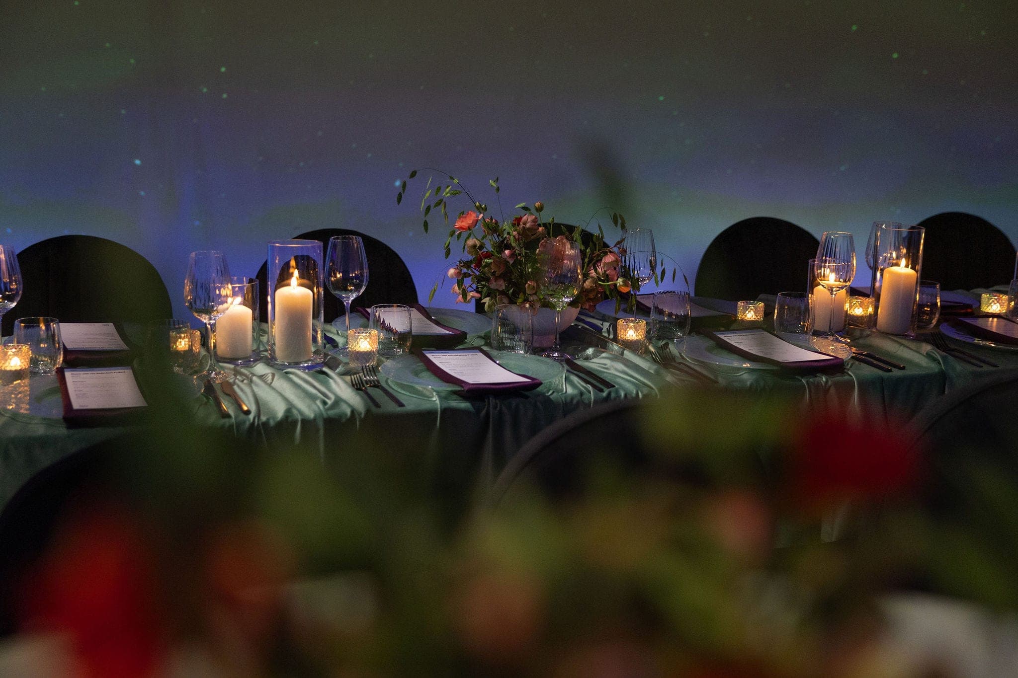 A candlelit dinner table set with crystal glassware, printed menus and a loose floral centrepiece, glowing against a wall lit like the northern lights