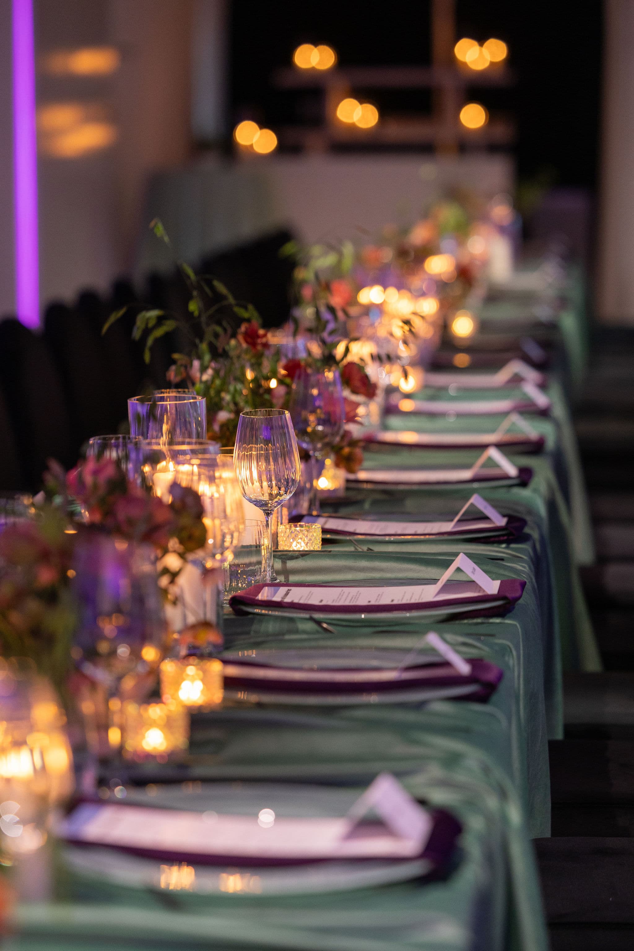 A long candlelit dinner table with sage linens, plum napkins, printed menus and low floral arrangements