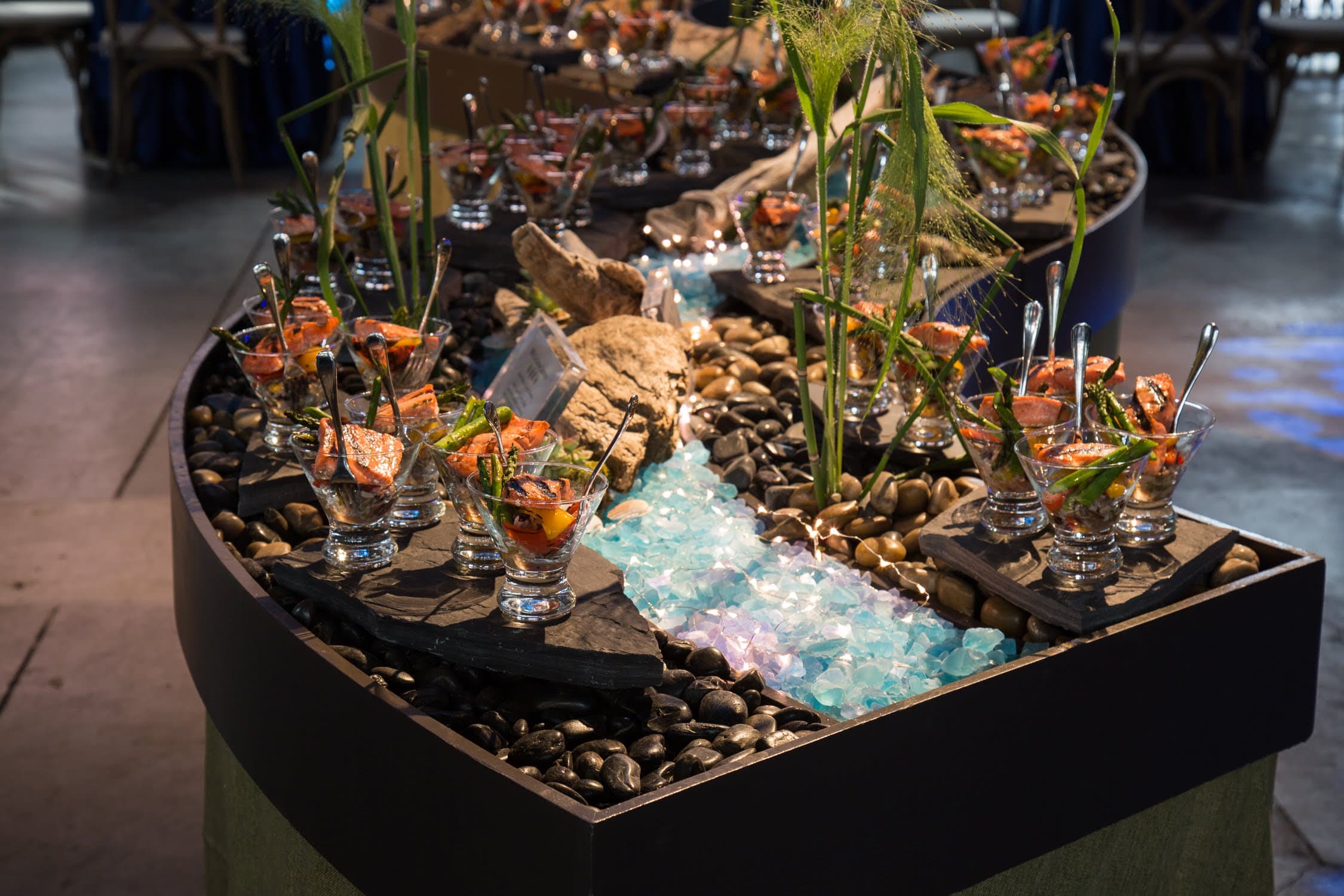A catering station of grilled salmon in glass coupes set on slate and river rock, with a lit sea-glass stream