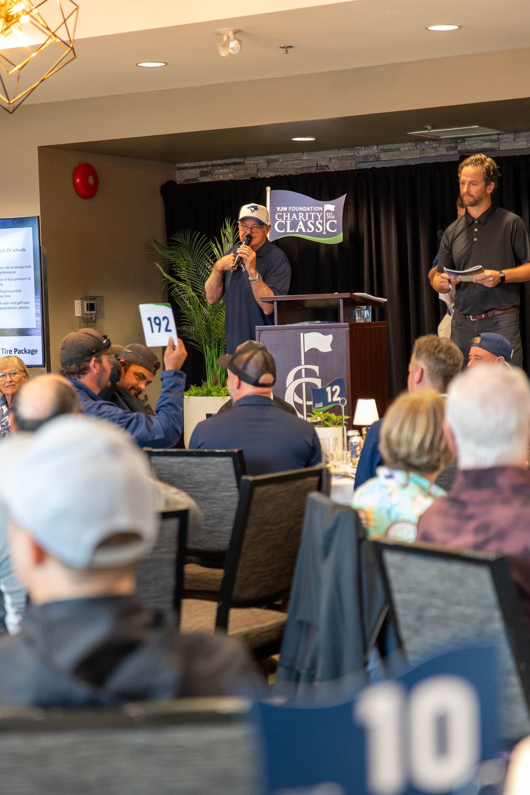 A live auction at a charity golf tournament dinner, with a guest raising bid paddle 192 toward the auctioneer at the podium