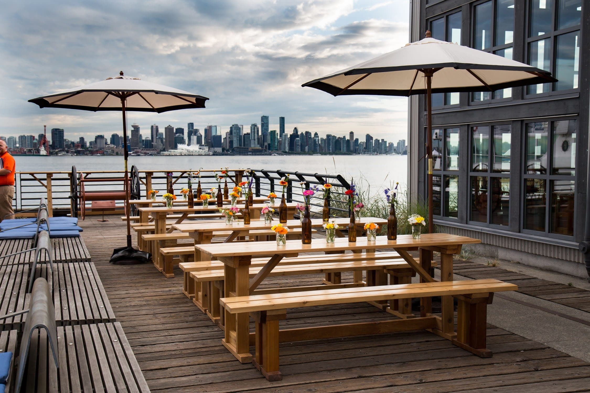 Cedar picnic tables with bottle vases of wildflowers on a waterfront deck, with the Vancouver skyline across the harbour