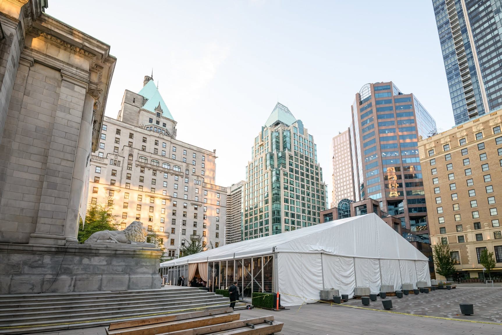 A white gala marquee set up on a downtown plaza beside stone steps and a lion statue, with city towers behind