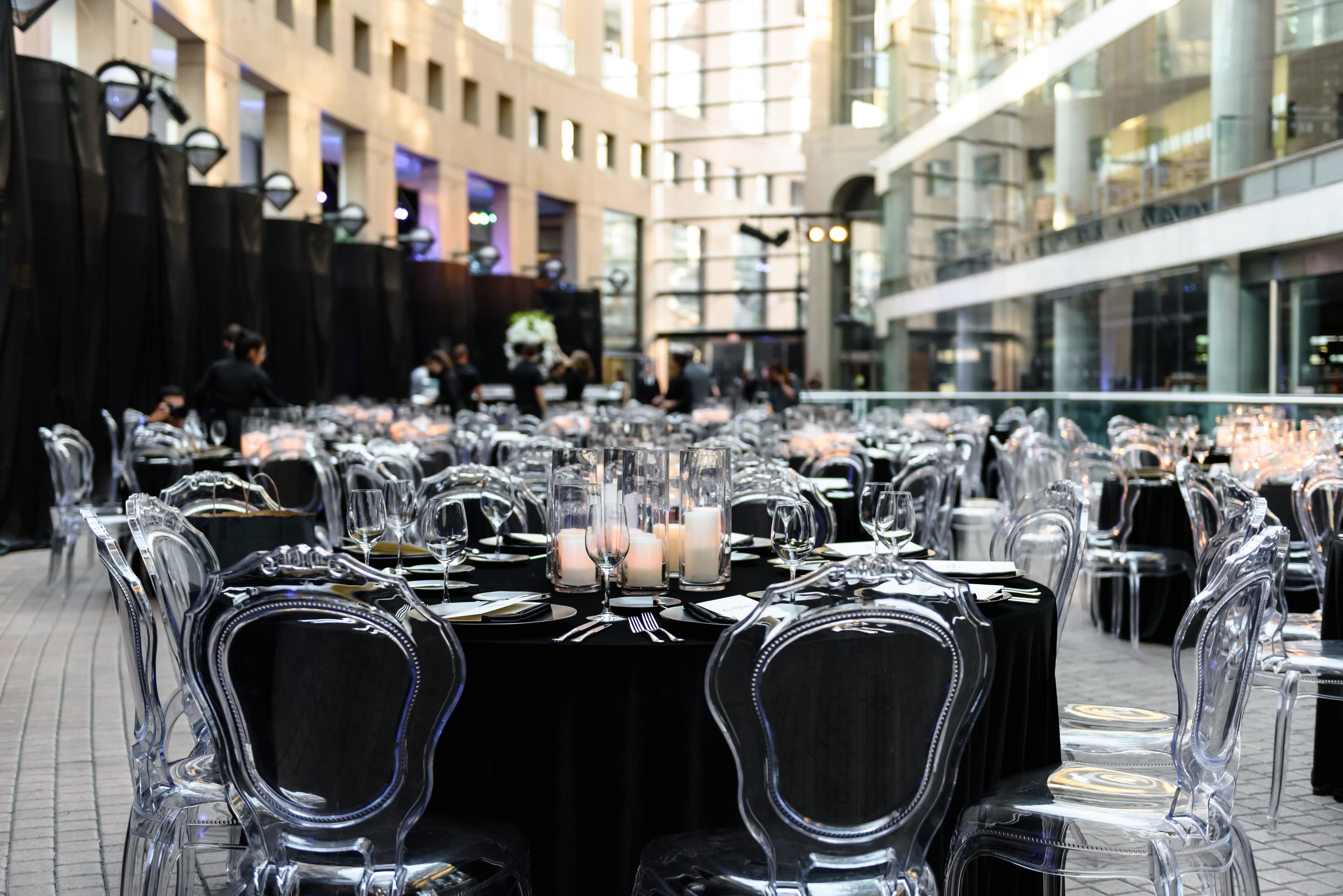Round dinner tables in black linen with clear ghost chairs and candlelit centrepieces in a stone atrium