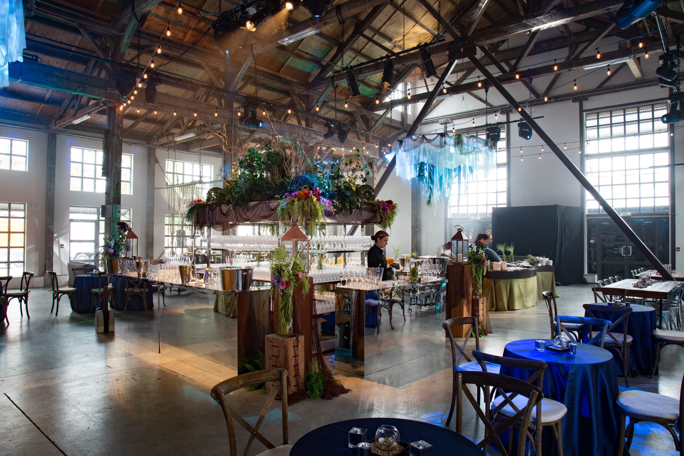 A warehouse venue set for a reception, with a mirrored bar under a suspended floral installation and string lights