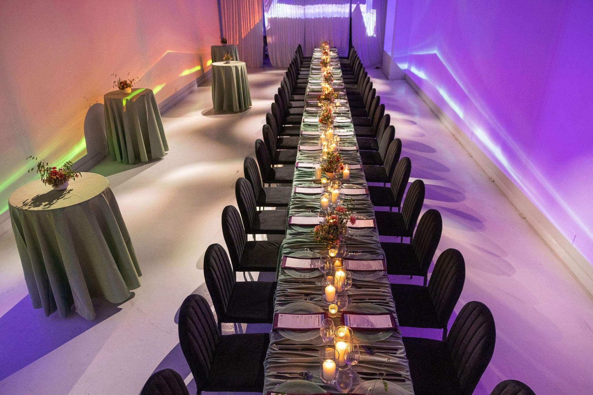 A long candlelit dinner table with sage linens and black chairs in a room washed with purple and green light